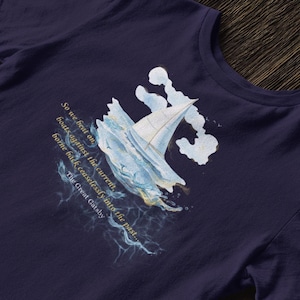 Puede incluir: Camiseta azul marino con una ilustraci&oacute;n en acuarela de un velero entre olas. El dise&ntilde;o incluye texto de "El Gran Gatsby": "So we beat on boats against the current, borne back ceaselessly into the past..."