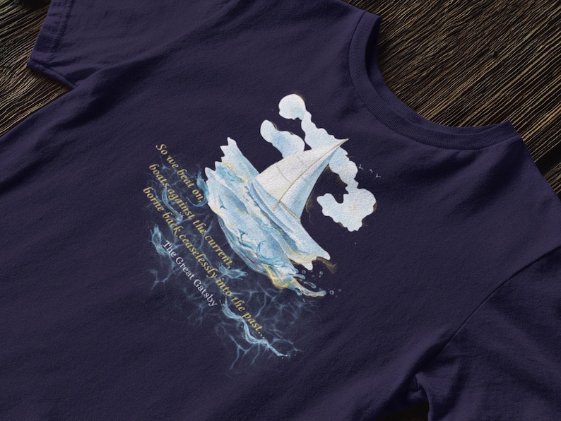 Puede incluir: Camiseta azul marino con una ilustraci&oacute;n en acuarela de un velero entre olas. El dise&ntilde;o incluye texto de "El Gran Gatsby": "So we beat on boats against the current, borne back ceaselessly into the past..."