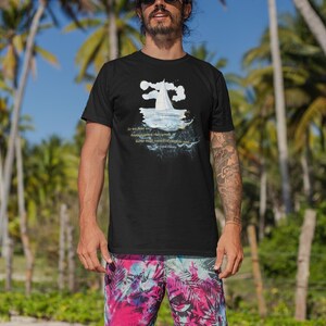 Puede incluir: Camiseta negra con un gr&aacute;fico de velero y texto de El Gran Gatsby. La persona lleva gafas de sol y pantalones cortos coloridos con estampado tropical. Palmeras y cielo azul en el fondo.