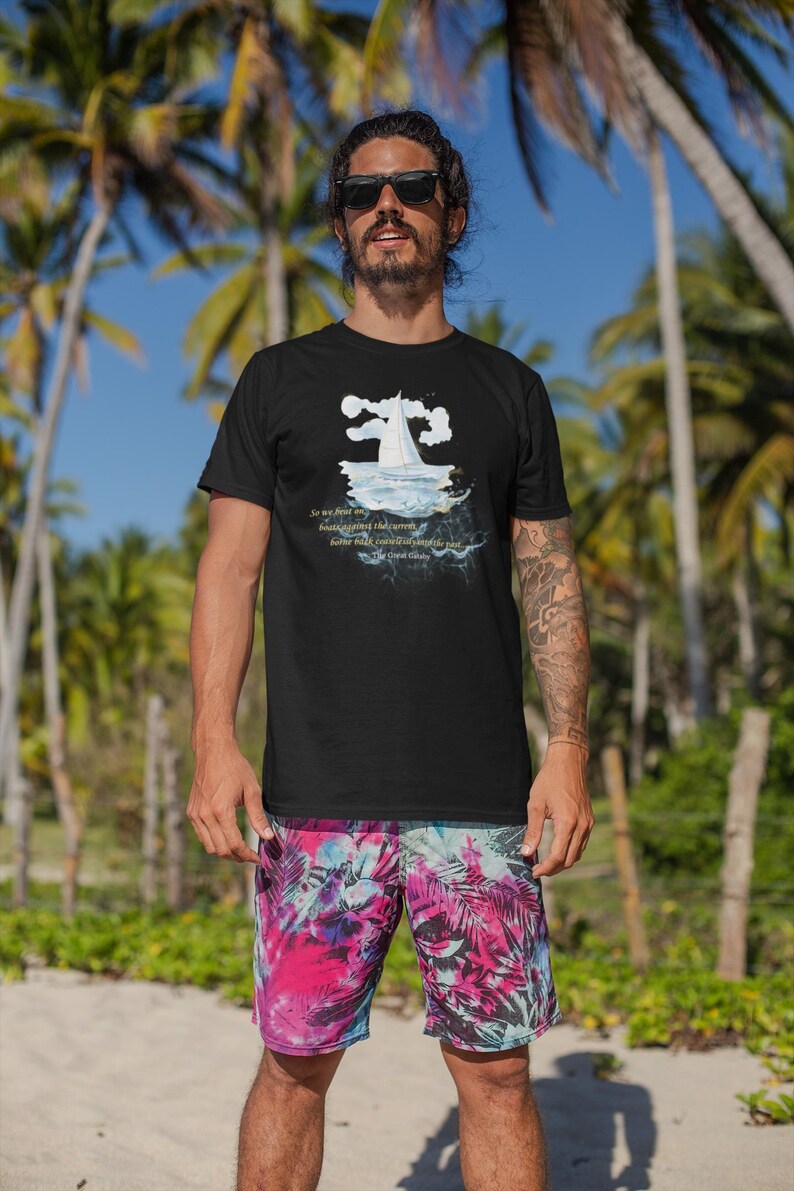 Puede incluir: Camiseta negra con un gr&aacute;fico de velero y texto de El Gran Gatsby. La persona lleva gafas de sol y pantalones cortos coloridos con estampado tropical. Palmeras y cielo azul en el fondo.