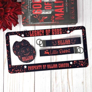 May include: A collection of merchandise featuring the book "God of Malice" by Rina Kent. Includes a license plate frame, keychains, and coasters with the text "Legacy of Gods", "Killian", and "Little Rabbit" in black and red.