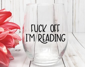 May include: A clear stemless glass tumbler featuring the black printed text FUCK OFF I'M READING centered on the front. The glass has a rounded base and a smooth rim, designed for holding beverages while maintaining a simple and functional shape.