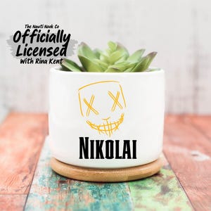 May include: White ceramic planter with a yellow graphic of a grinning face and the name "NIKOLAI" in black. The planter holds a green succulent and sits on a wooden coaster. The text "The Nauti Nook Co Officially Licensed With Rina Kent" is at the top.
