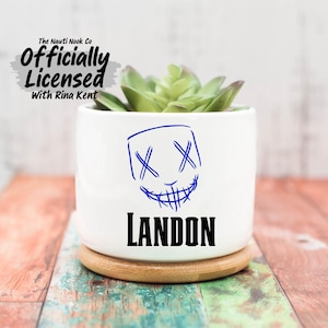 May include: White ceramic planter with a blue sketched mask design and the name "Landon" in black. The planter holds a green succulent and sits on a wooden coaster. The text "The Nauti Nook Co. Officially Licensed with Rina Kent" is at the top.