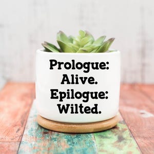 May include: A white ceramic pot with the text "Prologue: Alive. Epilogue: Wilted." contains a green succulent plant. The pot sits on a small, round wooden coaster. The background is a wooden surface.