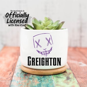 May include: White ceramic planter with a purple face design and the name "GREIGHTON" in black. The planter holds a green succulent and sits on a wooden coaster. The text "The Nauti Nook Co Officially Licensed With Rina Kent" is at the top.
