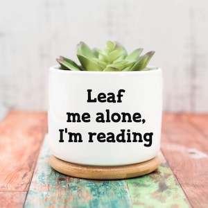 May include: A white ceramic planter with a green succulent plant. The planter has the text "Leaf me alone, I'm reading" in black. The planter sits on a small wooden coaster, with a colorful wooden background.