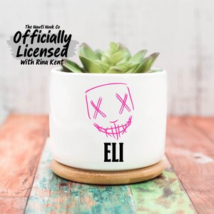 May include: A white ceramic planter with a pink, sketched face design and the name "ELI" in black. The planter holds a green succulent and sits on a wooden coaster. The text "The Nauti Nook Co Officially Licensed With Rina Kent" is at the top.