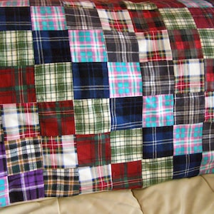 May include: A patchwork pillow with a variety of plaid patterns in red, green, blue, black, and purple. The pillow is made of squares of fabric sewn together, creating a cozy and colorful design. The pillow is resting on a tan surface.