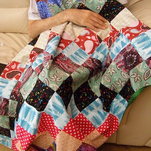 May include: A patchwork quilt with a variety of patterned squares in red, blue, green, black, and beige. The quilt features paisley, polka dots, musical notes, and floral designs. The quilt is folded over a person.