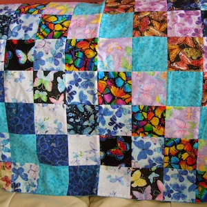 May include: A quilt made of colorful fabric squares featuring a butterfly pattern. The quilt is made up of various shades of blue, purple, and white, with accents of yellow, orange, and red. The butterflies are depicted in a variety of sizes and positions.