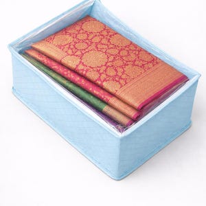 Op de afbeelding: Een lichtblauwe stoffen opbergdoos met gevouwen textiel. De textiel omvat een rood-goudkleurige stof, een groene stof en een paarse stof. De doos heeft een gewatteerd patroon.