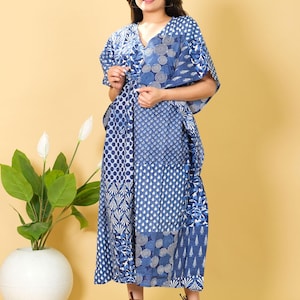 Può includere: Un lungo abito blu e bianco fantasia con scollo a V. L'abito presenta un design patchwork con vari motivi geometrici e floreali. La modella indossa sandali marroni. Una pianta in vaso bianca è sullo sfondo.