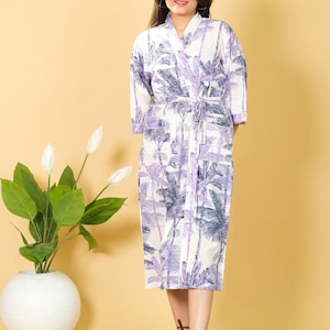 May include: A white robe with a palm tree print in shades of purple and blue. The robe has a wrap-around design with a matching belt. The robe is knee-length and has three-quarter sleeves. The model is wearing brown sandals.