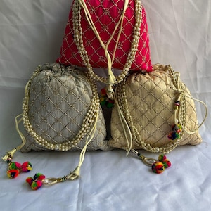 Pode incluir: Três sacos decorativos com cordão nas cores vermelho, dourado e prateado. Cada saco apresenta um padrão acolchoado e é adornado com um cordão de contas peroladas e detalhes de borlas coloridas. Os sacos estão sobre uma superfície branca.