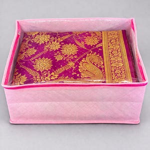 May include: A pink quilted storage box with a clear top, containing a magenta textile with gold floral patterns. The textile has a gold border and is neatly folded inside the box. The box is closed with a pink zip.