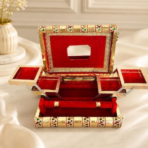 May include: A rectangular jewelry box with a gold-toned exterior featuring floral patterns and a red velvet interior. The box has a hinged lid with a central mirror, two tiered side compartments that extend outward, and a ring holder at the base.