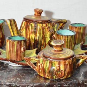 Può includere: Un servizio da tè in ceramica con un design a tronco d'albero. Il set comprende una teiera, una zuccheriera e tazze con piattini a forma di foglia. I colori sono marrone, giallo e verde, con interno turchese.