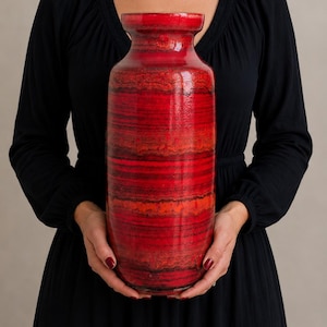 May include: A tall cylindrical ceramic vase featuring a vibrant red and orange gradient glaze with horizontal dark brown and black textured bands. The vase has a smooth, glossy finish, a slightly flared rim, and a rounded base, showcasing a rich color palette.