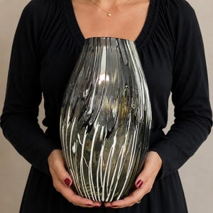 May include: A glass vase featuring a dark, smoky gray translucent base with a decorative pattern of vertical, flowing white and black streaks that wrap around the exterior surface, creating a textured, layered appearance on the rounded vessel.