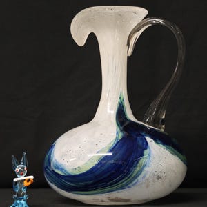 Può includere: Una grande brocca di vetro bianco con un manico trasparente e una parte superiore svasata. La brocca presenta accenti a spirale blu e verdi. Una piccola statuina di vetro blu di un coniglio che tiene una pergamena è accanto alla brocca.