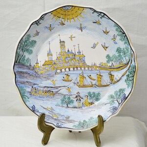 Può includere: Un piatto decorativo in ceramica con bordo smerlato. Il piatto presenta una scena dipinta a mano di una città sull'acqua, con barche, figure e uccelli. La tavolozza dei colori comprende blu, giallo e verde.