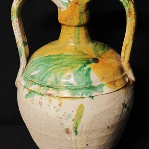 Puede incluir: Una jarra de cerámica antigua grande con dos asas. La jarra presenta una base de color crema con detalles en amarillo y verde en la parte superior y las asas. La jarra tiene un cuerpo bulboso y un cuello estrecho.