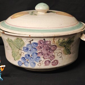 Può includere: Una pirofila in ceramica con coperchio, con un disegno dipinto a mano di uva nei toni del blu e del viola. Il piatto ha una base chiara con un bordo verde e rosa sul coperchio. Le maniglie sono a forma di foglia.
