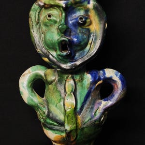 May include: A whimsical ceramic sculpture of a figure with a face, hat, and arms. The sculpture is painted in shades of green, blue, and yellow. The figure has an open mouth and appears to be surprised.
