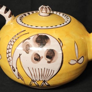 Può includere: Una teiera in ceramica gialla con corpo arrotondato, manico curvo e beccuccio. La teiera presenta un disegno dipinto di una creatura bianca e marrone sul lato. Il manico e il pomello del coperchio hanno strisce marroni e bianche.