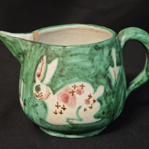 Può includere: Un piccolo brocca in ceramica verde con un disegno di coniglio bianco e accenti rosa. La brocca ha un beccuccio e un manico. Il coniglio ha macchie rosa e segni marroni a forma di croce. Lo sfondo è verde screziato.