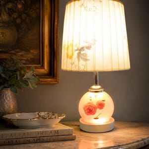 Può includere: Una lampada da tavolo decorativa con un design floreale. La lampada ha una base rotonda in vetro smerigliato con rose rosa dipinte. Il paralume è color crema con un design a pieghe e accenti floreali. La lampada è su un tavolo con piano in marmo.