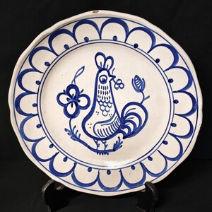 Può includere: Piatto in ceramica bianca con un disegno di gallo blu al centro. Il piatto ha un bordo smerlato e un bordo floreale blu. Il gallo tiene un fiore nel becco. Diametro di circa 25 cm.