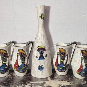 Può includere: Un set di pezzi da portata in ceramica vintage. Il set comprende un vaso alto e strutturato e quattro brocche. Le brocche e il vaso sono decorati con disegni colorati e astratti con tamburi e figure. I manici delle brocche hanno un motivo a pois neri e gialli.