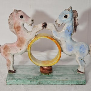 Può includere: Scultura in ceramica raffigurante due cavalli stilizzati, uno rosa e marrone, l'altro blu, che si fronteggiano e sostengono un anello giallo. La base è rettangolare con effetto marmorizzato verde e bianco. Oggetto decorativo.