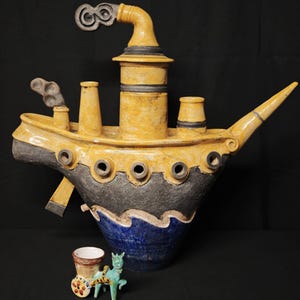 Può includere: Una grande scultura artigianale in ceramica a forma di barca con colori giallo, nero e blu. La barca ha un camino, oblò e un beccuccio decorativo. Un piccolo carretto trainato da cavalli turchese è di fronte alla barca.