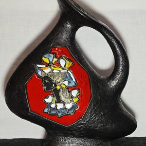 Può includere: Brocca in ceramica nera con superficie strutturata e forma unica. Presenta un inserto rosso ottagonale con il profilo di una donna con cappello, accentuato da dettagli gialli, blu e bianchi. La brocca ha un manico e un beccuccio.