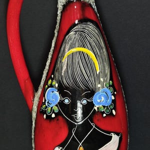 Può includere: Un vaso in ceramica rosso con un disegno di volto nero, capelli bianchi e orecchini floreali blu. Il vaso ha un manico rosso e un collo strutturato grigio. Sono visibili anche una fascia gialla e un ciondolo.