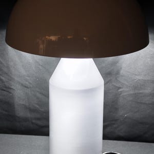 May include: A modern table lamp with a brown, dome-shaped lampshade and a white, cylindrical base. The lamp emits a soft, diffused light. The base is approximately 10 inches tall.