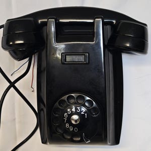 Può includere: Un telefono nero vintage con quadrante rotante e una cornetta su ciascun lato. Il telefono ha un tastierino rettangolare e un quadrante circolare con numeri. Il telefono è montato a parete e ha fili che si estendono dalla parte inferiore.