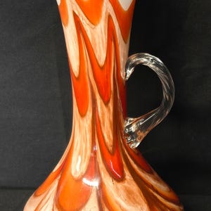 May include: A tall, decorative glass pitcher with a white top and a clear, twisted handle. The pitcher features a vibrant pattern of orange and cream-colored swirls. The pitcher has a spout for pouring.