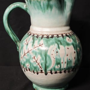Può includere: Brocca in ceramica con corpo arrotondato e manico. La brocca presenta una base bianca con dettagli dipinti in verde e marrone, tra cui un bordo decorativo e figure stilizzate. La parte superiore e il manico sono di colore verde pieno.