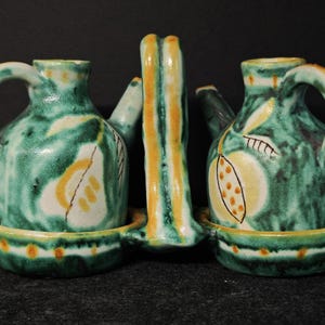 Può includere: Un set di sale e pepe in ceramica con una maniglia centrale. Il set presenta due piccoli brocche con manici e beccucci, dipinti in verde con accenti gialli e bianchi. Il set poggia su un vassoio abbinato.