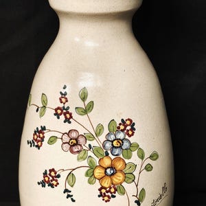 Può includere: Vaso in ceramica color crema con design floreale dipinto a mano. Il vaso presenta una parte superiore svasata con finiture marroni e verdi. Il design floreale include fiori gialli, blu, rosa e rossi, con foglie verdi. La parola "Santoriello" è scritta sul vaso.