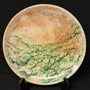 Può includere: Un piatto rotondo in ceramica con un design unico. Il piatto presenta una miscela di colori, tra cui pesca, beige e verde, creando un paesaggio astratto. Le aree verdi assomigliano a fogliame, mentre il pesca e il beige suggeriscono un tramonto.
