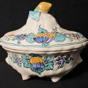May include: A cream-colored ceramic serving dish with a lid, adorned with hand-painted floral designs in blue, yellow, and teal. The dish has a unique shape with a small handle on the lid and sits on three small feet.