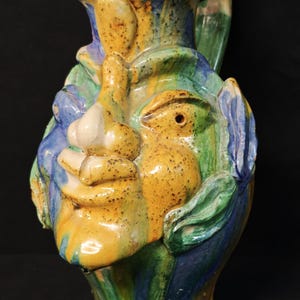 Può includere: Brocca in ceramica con design a volto. La brocca ha un manico ed è dipinta con colori giallo, verde, blu e bianco. Il volto ha naso, occhi e bocca prominenti. La brocca è un'opera d'arte unica.