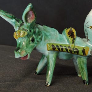 May include: A glazed ceramic donkey figurine in shades of green, featuring a harness with black markings and a saddle. The donkey is attached to a cart component with a white base, decorated with a painted brown and yellow floral design.
