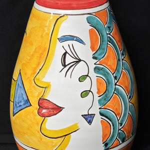 Può includere: Vaso in ceramica dipinto a mano con un design stilizzato del viso. Il vaso presenta una parte superiore rossa, una base arancione e un viso giallo e bianco con accenti blu, verdi e arancioni. Il design ricorda lo stile artistico di Picasso.