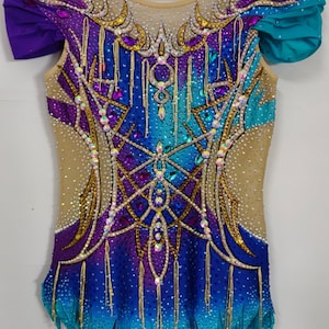 Puede incluir: Maillot adornado con joyas de colores vibrantes, con un degradado de tonos azules y morados. La prenda presenta intrincados adornos dorados y de cristal, con toques de color turquesa y morado en las mangas. El texto "FROZENFANTASIA" está impreso en la parte inferior.